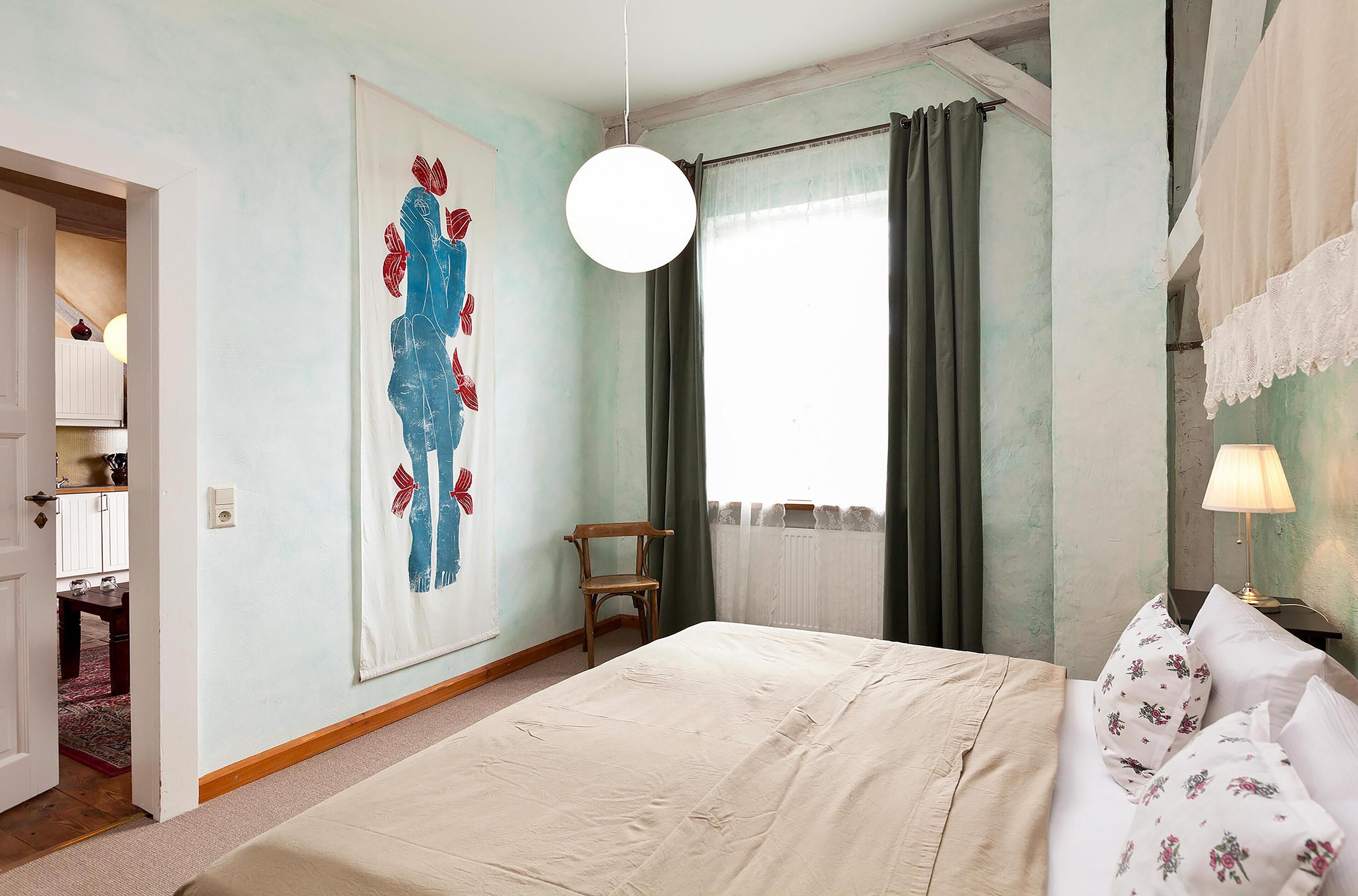 A bedroom with a large bed, a hanging light fixture, a chair, and a painting on the wall.