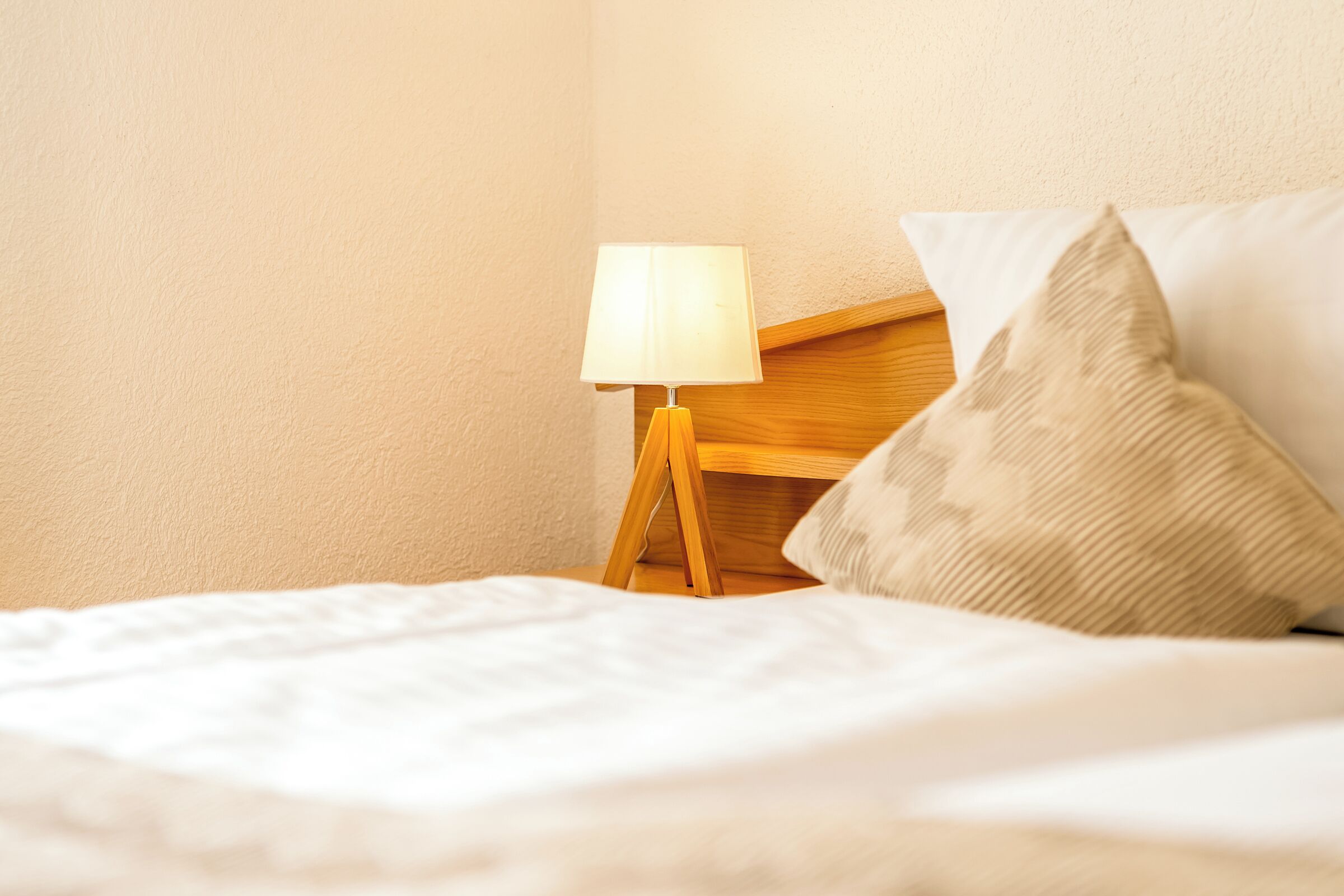A neatly made bed with a white comforter, a wooden bedside table with a lamp, and a textured pillow.