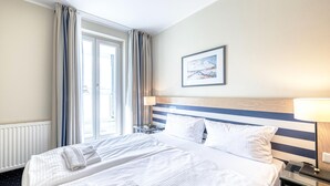 Ein Hotelzimmer mit zwei Betten, einem großen Fenster mit Vorhängen, einem Nachttisch mit Lampe sowie einem gerahmten Bild an der Wand.