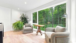 Ein modernes Wohnzimmer mit zwei Sofas, einem Holztisch und großen Fenstern mit Blick auf einen Wald.