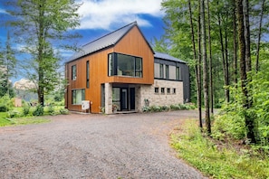 A modern two-story house with a stone facade, large windows, and a gravel driveway. - Spa & River | Le Saint-Gabriel (Saint-Gabriel-de-Valcartier)