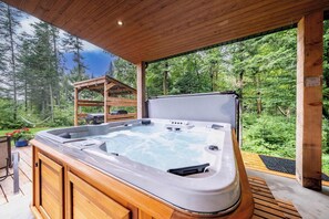 A hot tub with a wooden deck and a pergola, surrounded by a forested area.