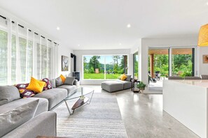A modern living room with a grey sofa, a glass coffee table, and a view of a lush garden through large windows.
