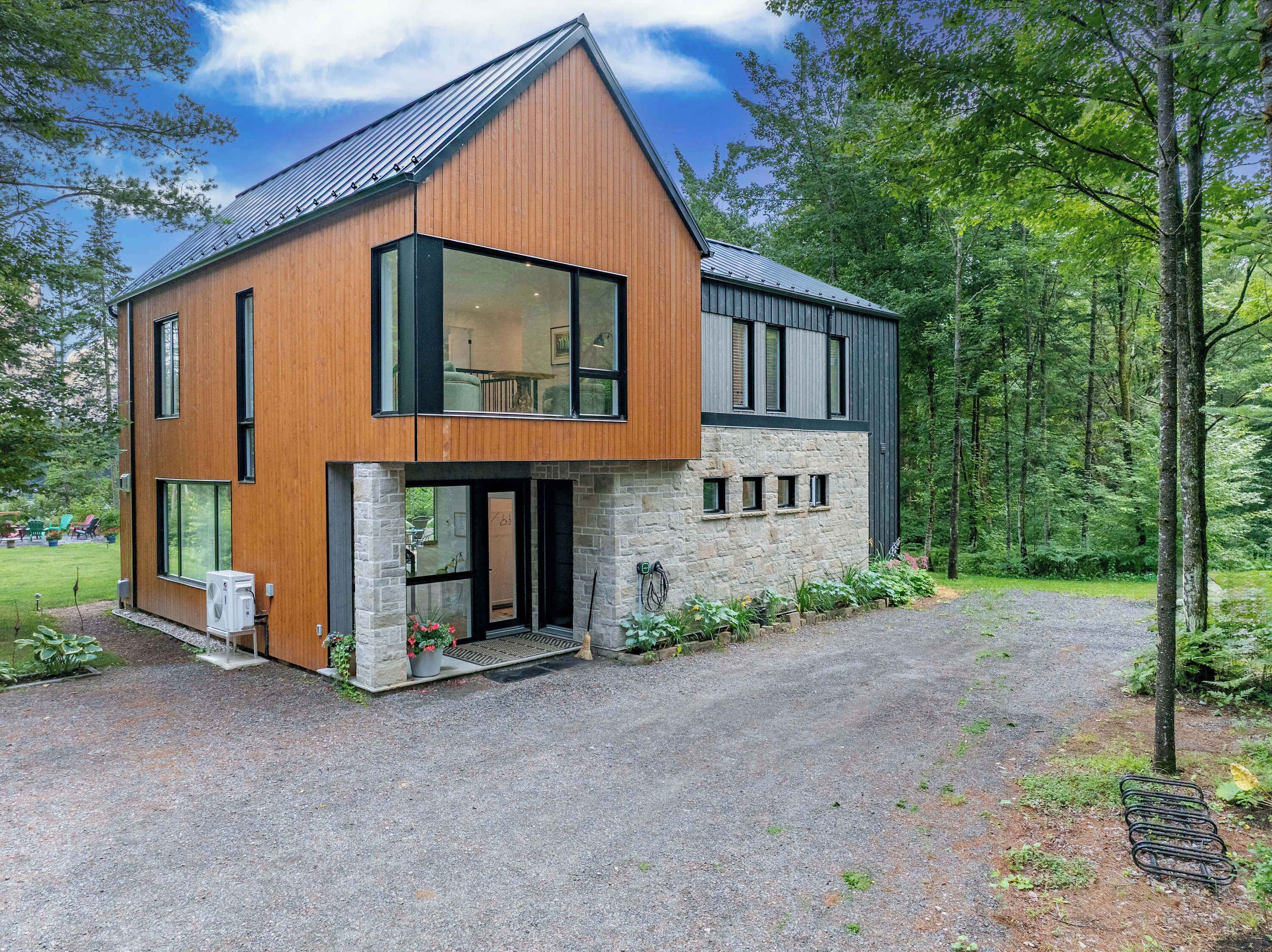 A modern two-story house with a stone foundation and wooden siding, a driveway, and a forested backdrop.