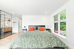 A modern bedroom with a large bed, a nightstand, a bedside table, a window with blinds, and a view of greenery outside. - Spa & River | Le Saint-Gabriel (Saint-Gabriel-de-Valcartier)