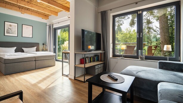 A modern living room with a sofa, a TV, and a dining table. There is a bookshelf with books and a window with a view of greenery.