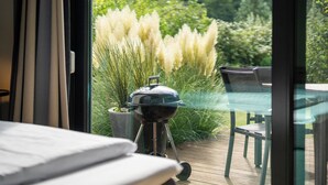 A modern outdoor dining area with a glass table and chairs, a barbecue grill, and a view of a lush garden with tall grasses.
