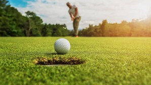 Uma bola de golfe num campo verde com um buraco, e uma pessoa ao fundo.