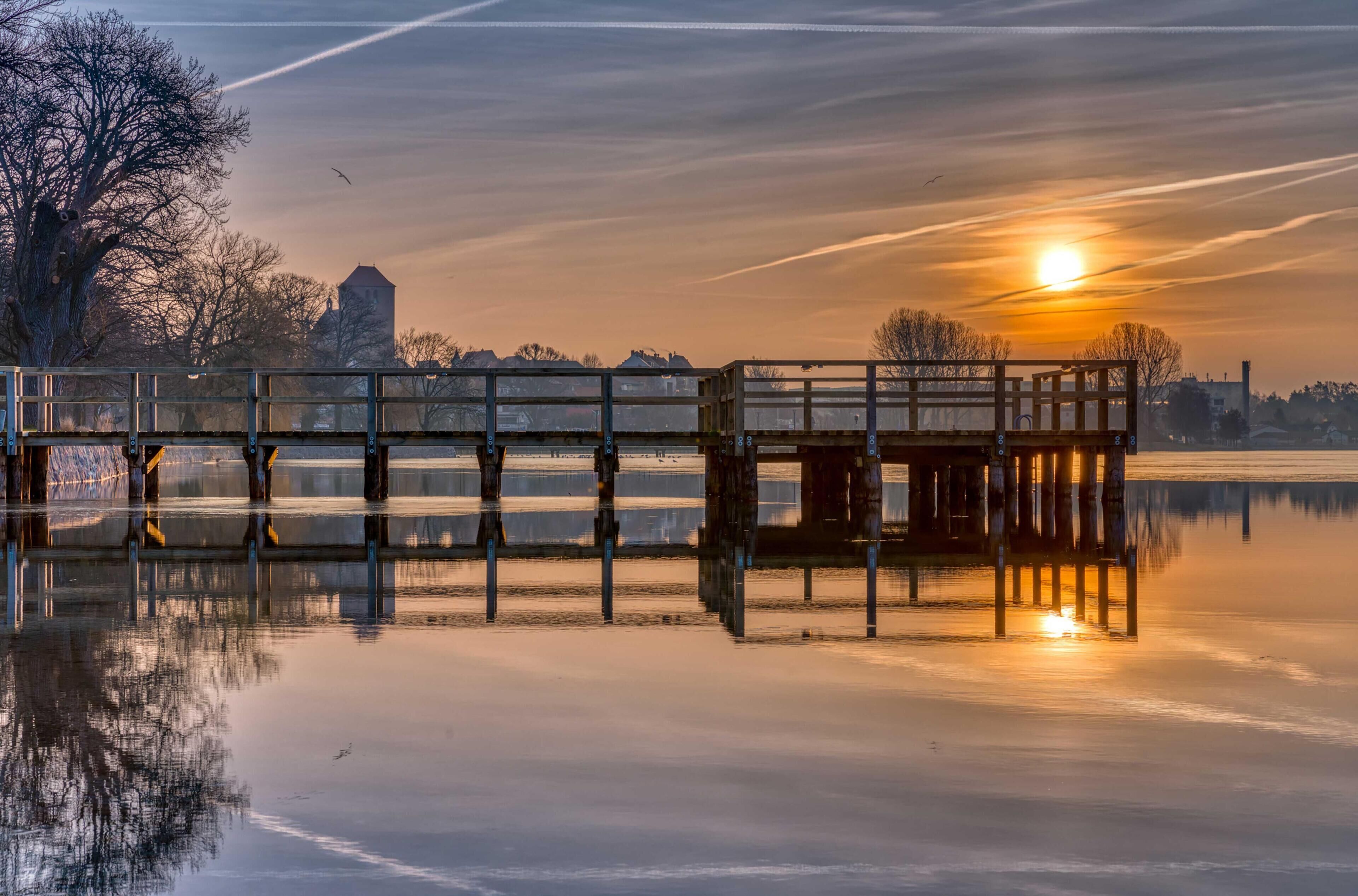 A wooden pier extending into a calm body of water, with a sunset in the background and a clear sky.