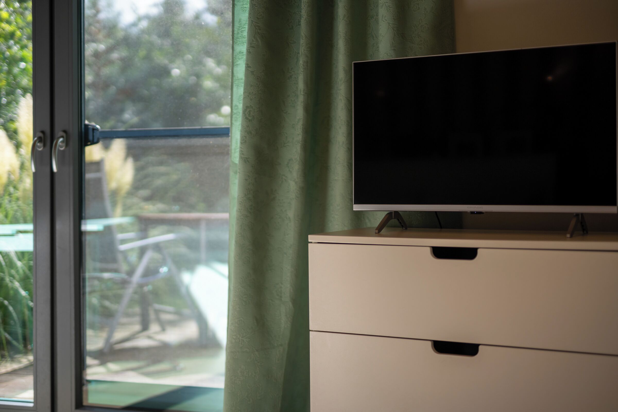 Una habitación con una televisión sobre un mueble y un ventanal corredera que da a un espacio exterior con vegetación.