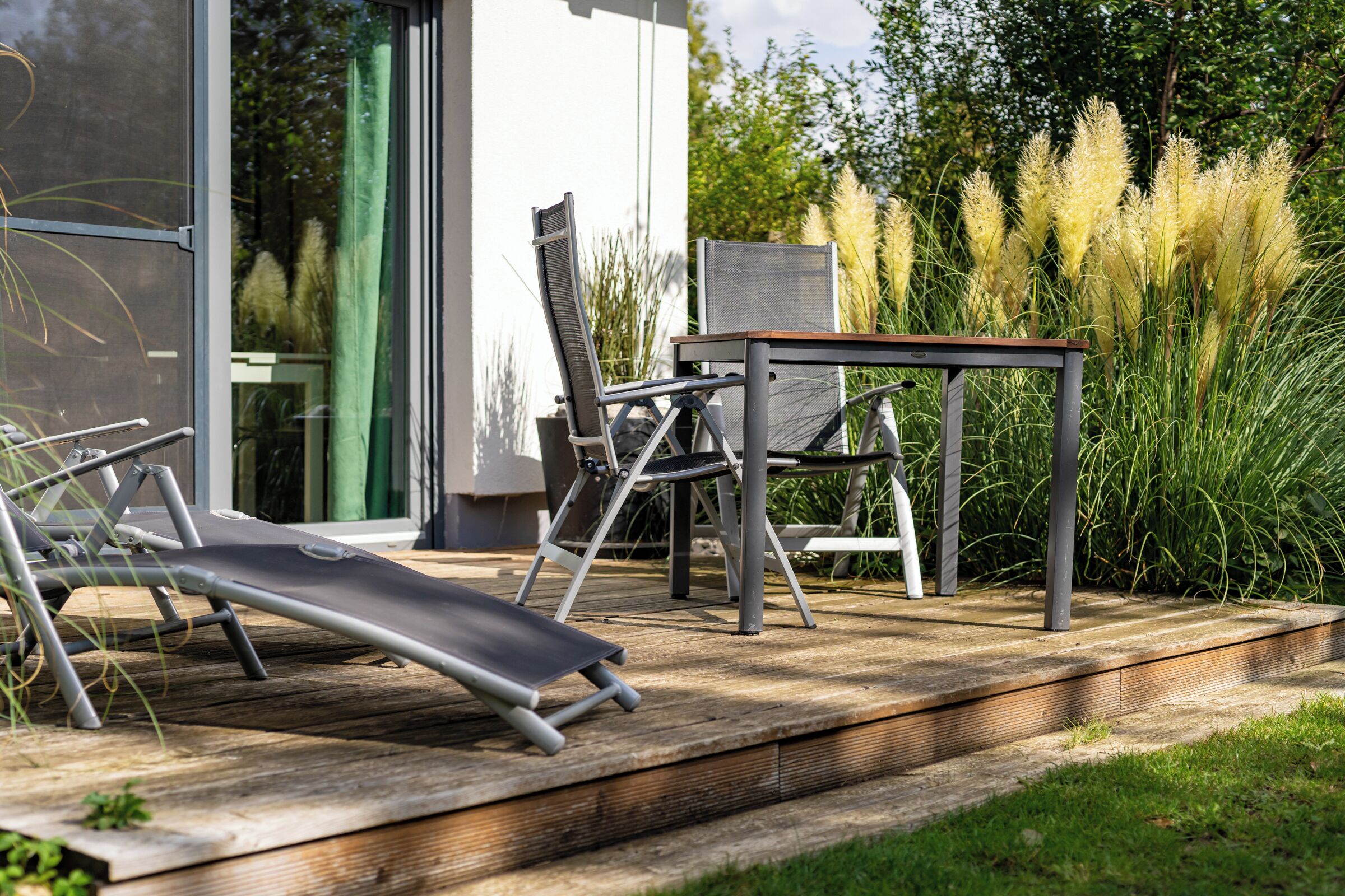 Eine Terrasse mit Tisch und Stühlen, umgeben von hohem Gras und einer Glastür.