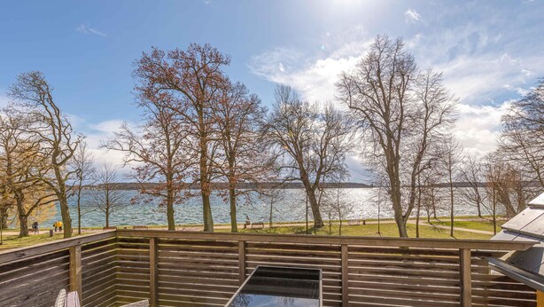 Une terrasse extérieure avec une table à plateau de verre et des chaises en osier, donnant sur un lac et une forêt.