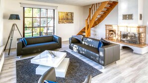 Una sala de estar moderna con dos sofás de cuero negro, una mesa de centro blanca y una chimenea. Una escalera de madera conduce a un nivel superior.