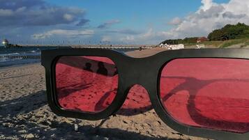 A pair of sunglasses on a sandy beach with a view of a pier and buildings in the background.