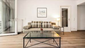 Un soggiorno con un tavolino in vetro, un divano con cuscini a quadri, un quadro appeso al muro e una porta che dà accesso a un'altra stanza.