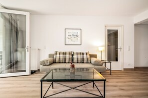 Uma sala de estar com uma mesa de centro de vidro, um sofá com almofadas xadrez, um quadro na parede e uma porta que dá para outro cômodo.