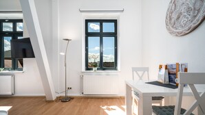 En moderne stue med et hvidt bord, en gulvlampe og et stort vindue med udsigt til himmel og skyer.