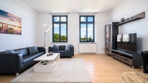 Ein modernes Wohnzimmer mit einem schwarzen Sofa, einem weißen Couchtisch und einem großen Flachbildfernseher. Zwei Fenster bieten Blick ins Grüne, und an der Wand hängt ein Gemälde.