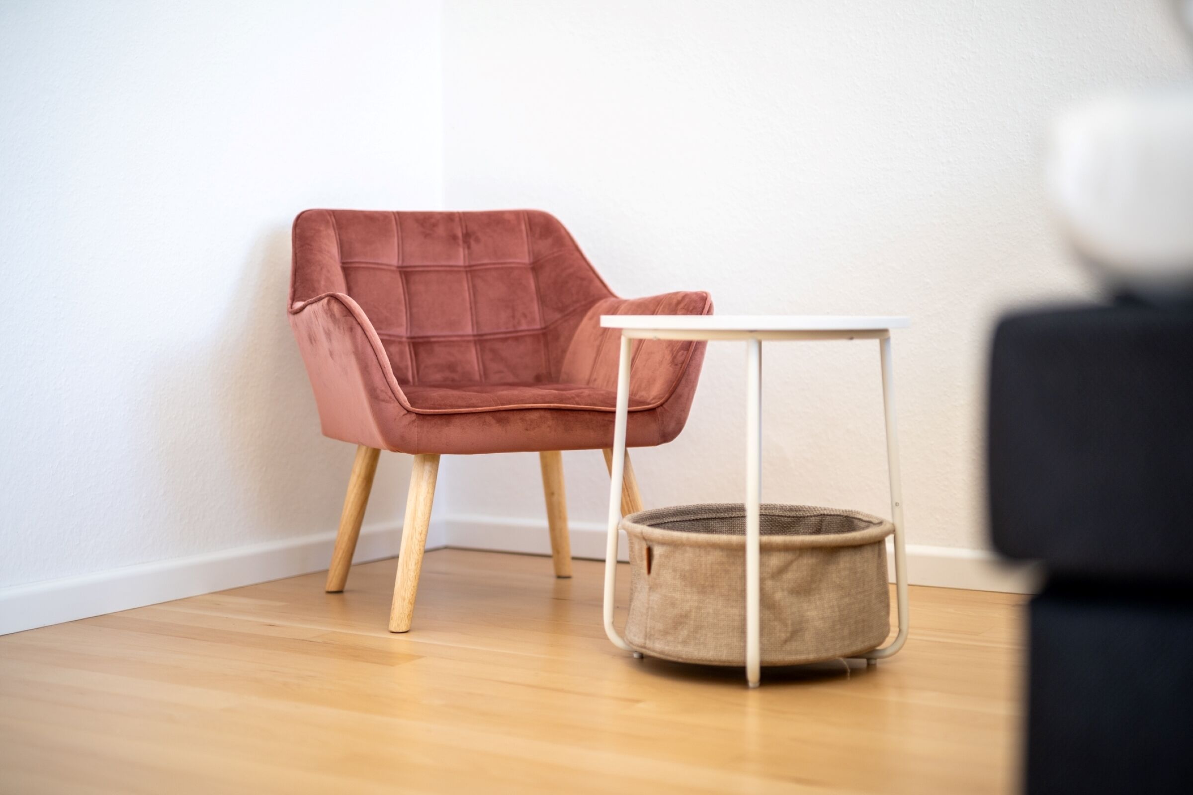 Una habitación con piso de madera, una silla rosa, una pequeña mesa blanca y una cesta redonda.