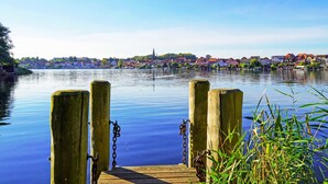 Eine Holzstege führt in ruhiges Wasser, im Hintergrund sieht man eine Stadt.