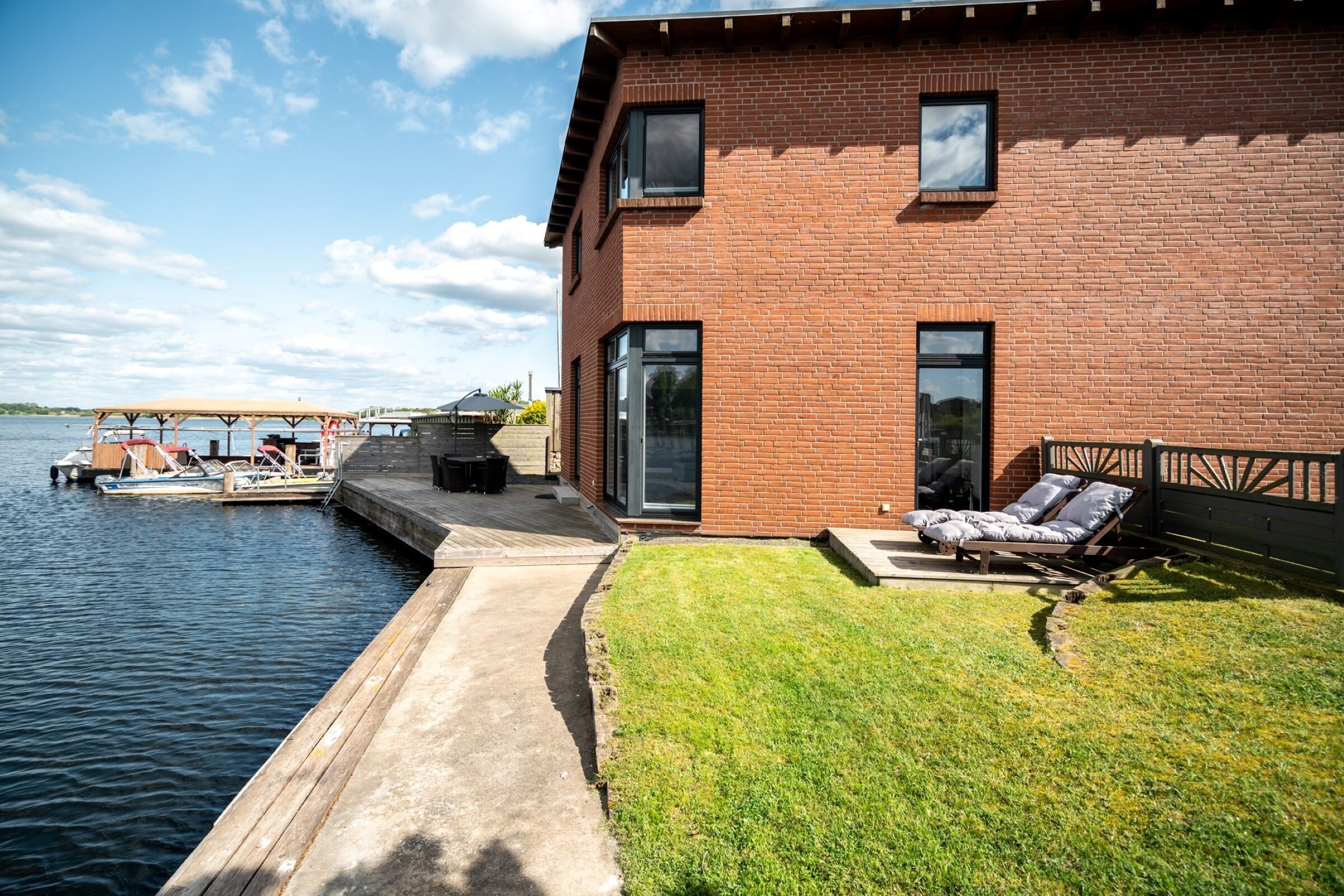 A modern house with a brick exterior, a deck by the water, and a lounge area with outdoor furniture.