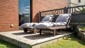 A wooden bed with wheels on a wooden deck, positioned in front of a brick building with a glass door.