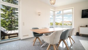 Un espace de restauration moderne avec une table et des chaises en bois, de grandes fenêtres offrant une vue sur les arbres et les bâtiments, et un lustre.