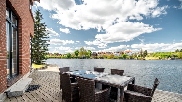 Une terrasse en bois avec une table à plateau de verre et des chaises en osier, donnant sur une étendue d’eau, avec des bâtiments en arrière-plan.