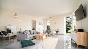 Un salon spacieux comprenant un canapé, une table basse et des fauteuils, un téléviseur fixé au mur et une grande fenêtre offrant une vue sur l’extérieur.