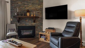 Ein Wohnzimmer mit Kamin, Flachbildfernseher, Ledersessel und einer Holzregal mit Büchern.