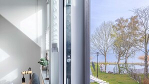 Une chambre moderne dotée d’une grande fenêtre, d’un luminaire suspendu et offrant une vue sur un lac avec une terrasse en bois.