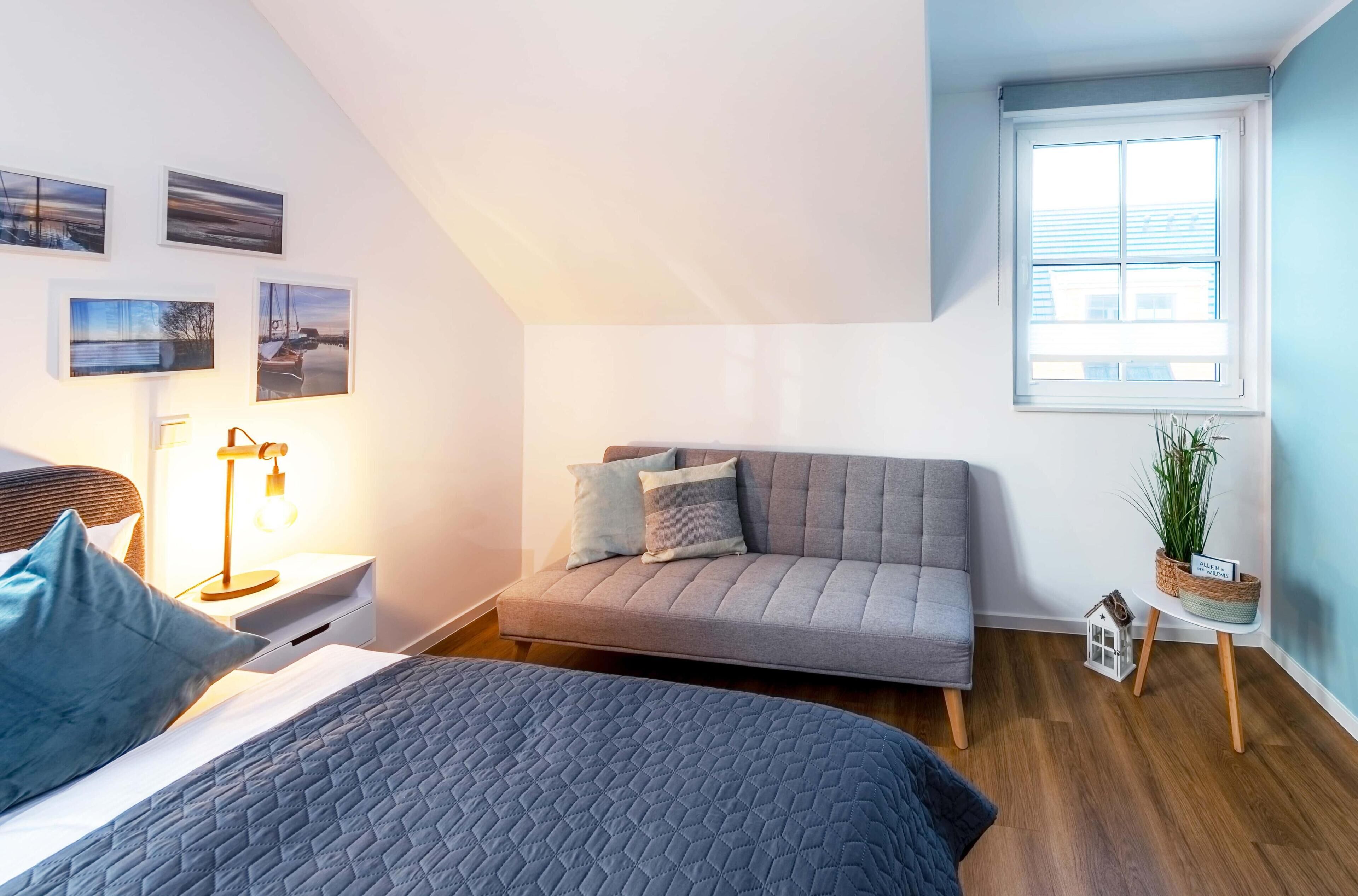 Et moderne soveværelse med en seng, en sofa, et natbord med lampe og et vindue med udsigt til bygninger.