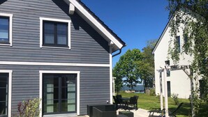 Une maison moderne à deux étages, avec une façade grise, de grandes fenêtres et un patio aménagé avec du mobilier d’extérieur.