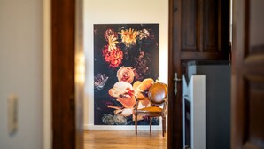 Un couloir au plancher de bois mène à une pièce où un grand tableau floral orne le mur, et une chaise se trouve au premier plan.