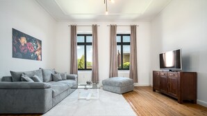 Un salon moderne avec un canapé gris, un pouf et une table basse en verre. On aperçoit un téléviseur posé sur un meuble en bois, et un tableau accroché au mur.