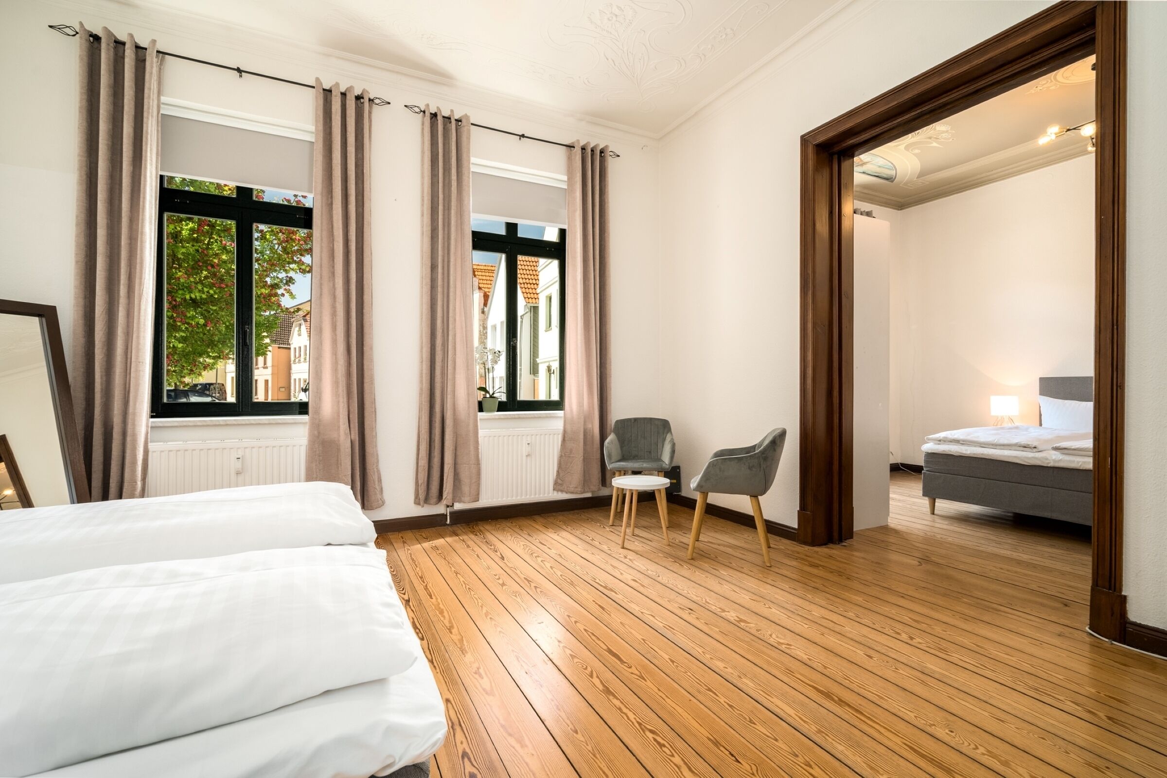 Ein Hotelzimmer mit Holzboden, einem Bett, einem Stuhl und Blick auf die Straße durch große Fenster.