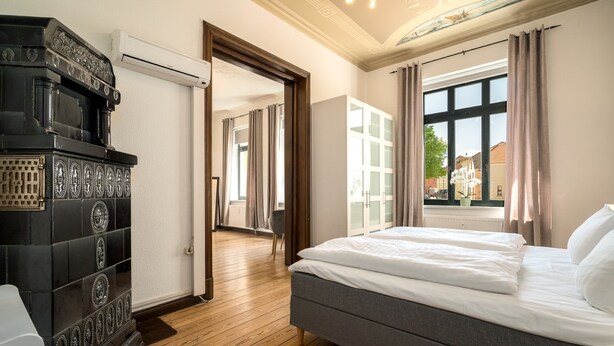Une chambre à coucher avec un lit, un plancher en bois, un poêle noir et une fenêtre avec des rideaux.