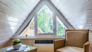 Un acogedor dormitorio en el ático, con techo inclinado, un ventanal con vistas a árboles, una silla de mimbre y una mesita con un jarrón de flores.