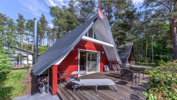 Una cabaña roja con techo inclinado, una terraza con una silla de lounge y una mesa, y una antena parabólica.