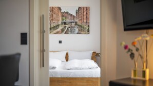 Habitación de hotel con una cama de madera, ropa de cama blanca, un cuadro de un paisaje urbano en la pared y un florero con flores en un tocador.