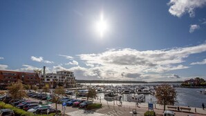 A marina with boats docked, a parking area with cars, and buildings in the background.