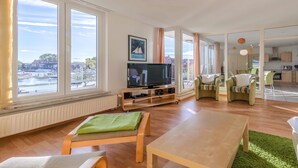 A spacious living room with a large TV, wooden furniture, and a view of the city through large windows.