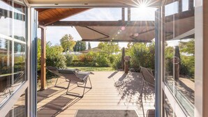Un espacio al aire libre techado con una terraza de madera, dos sillones de descanso y una pérgola que da sombra.