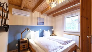 Un dormitorio con techo de madera, cama con ropa de cama blanca, mesita de noche con lámpara y ventana con vistas al verdor.