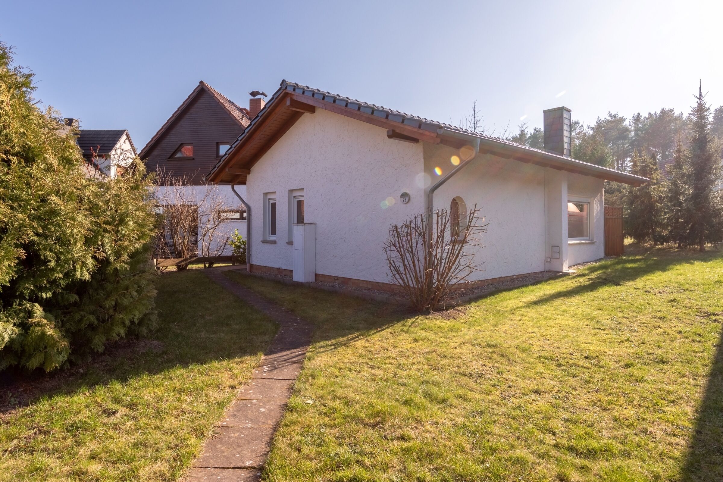 Une maison de plain-pied avec un toit en pente, une cheminée bien visible et un petit jardin devant, avec un chemin menant à l’entrée.