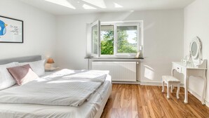 Ein Schlafzimmer mit einem Bett, einem Schreibtisch, einem Stuhl, einem Heizkörper und einem Fenster mit Blick auf Grünfläche.