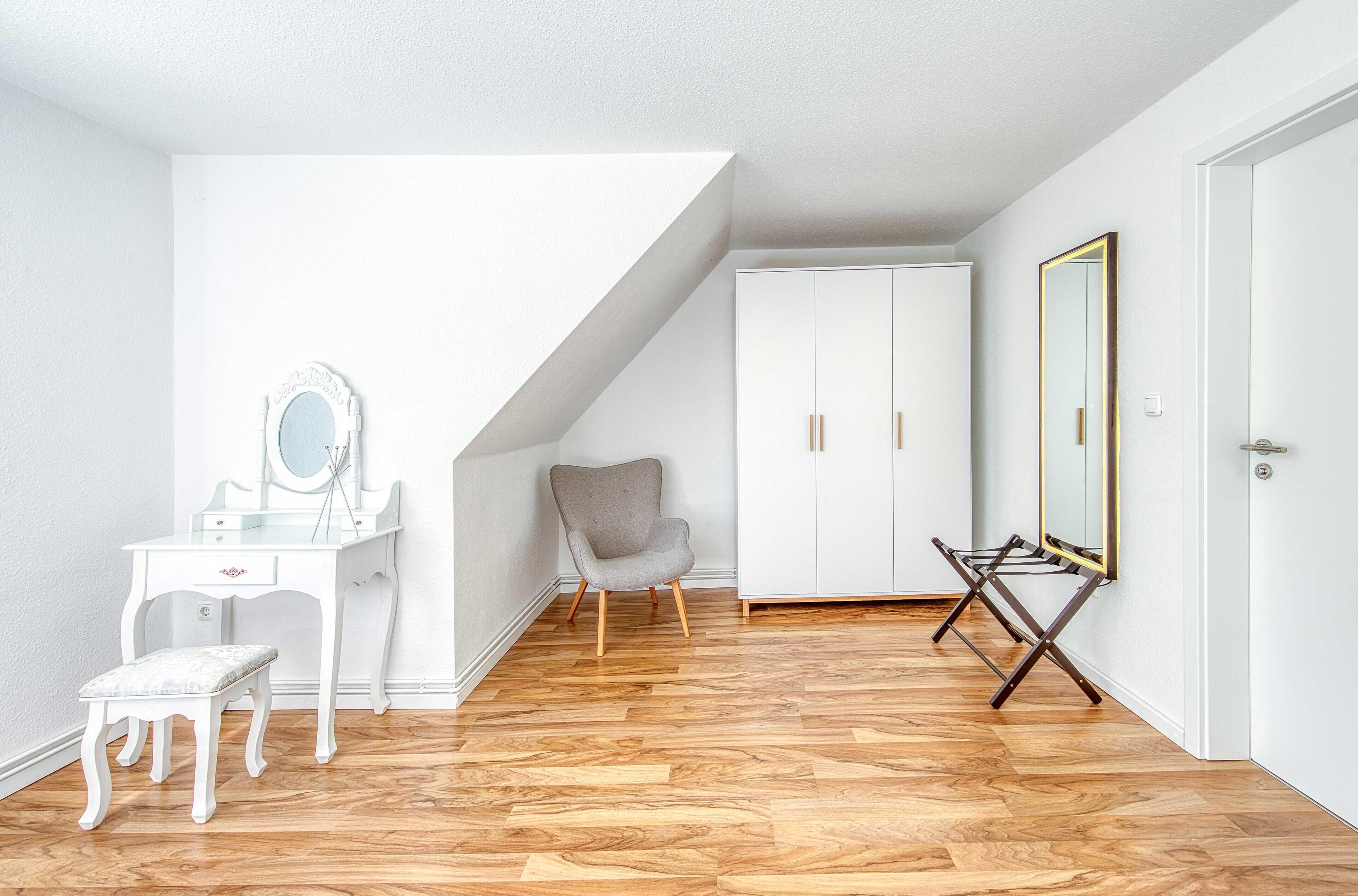 Ein Zimmer mit weißem Schminktisch, einem Stuhl, einem Kleiderschrank und einem großen Spiegel.