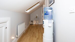 Un couloir doté d’un plancher en bois, de murs blancs, d’un radiateur, d’un distributeur d’eau et d’une petite table avec une lampe.