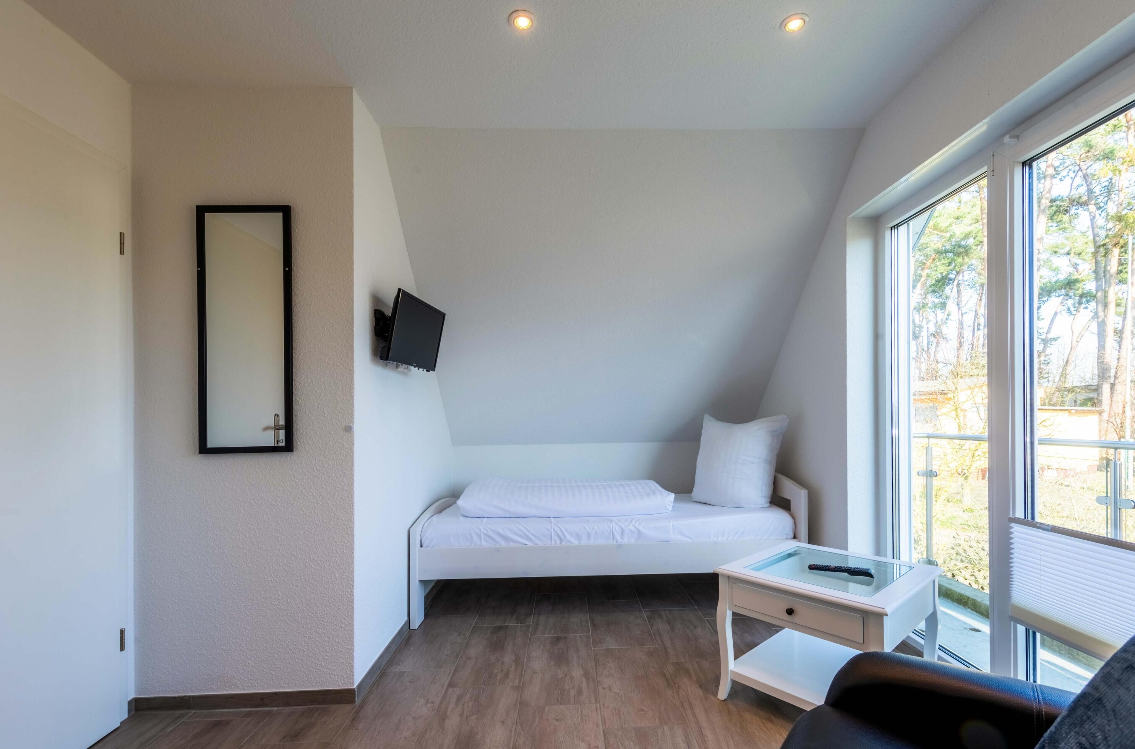 A modern bedroom with a bed, a TV mounted on the wall, a small table, and a window with a view of trees.