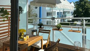 Una terraza de madera con una mesa y sillas, plantas en macetas y vistas a un edificio con contraventanas azules.